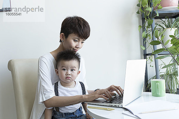 Mutter mit süßem Jungen auf dem Schoß arbeitet über Laptop zu Hause