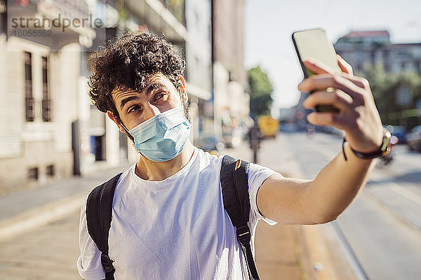 Nahaufnahme eines jungen Mannes mit Maske  der ein Selfie mit seinem Smartphone macht  während er in der Stadt steht