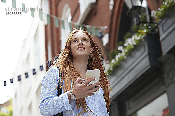 Lächelnde rothaarige Frau schaut weg  während sie mit ihrem Smartphone gegen ein Gebäude in der Stadt läuft