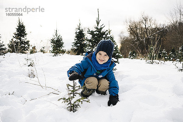 Lächelnder Junge zeigt Baum  während er auf schneebedecktem Land vor dem Himmel kauert