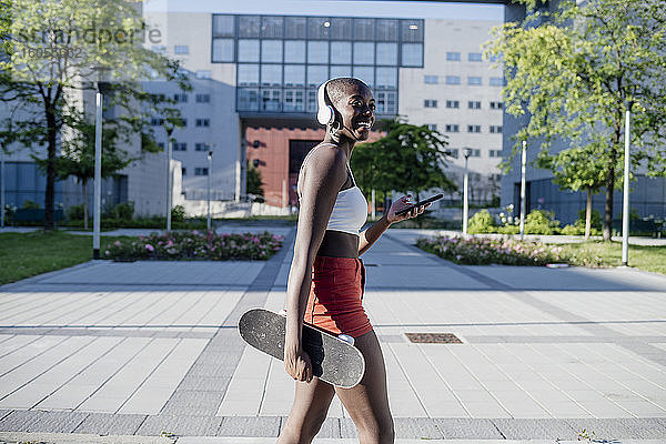 Glückliche Frau  die ein Skateboard hält und ein Mobiltelefon benutzt  während sie auf einem Fußweg in der Stadt spazieren geht
