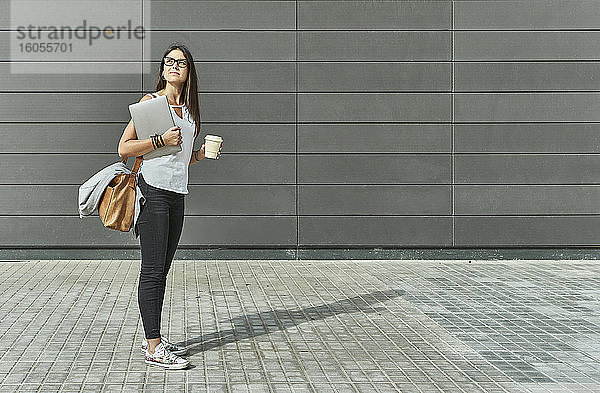 Nachdenkliche Geschäftsfrau  die einen Laptop und ein Einwegglas hält  während sie auf der Straße steht