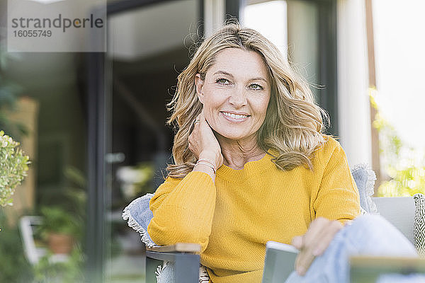 Porträt einer lächelnden reifen Frau  die mit einem digitalen Tablet auf einer Terrasse sitzt und in die Ferne schaut