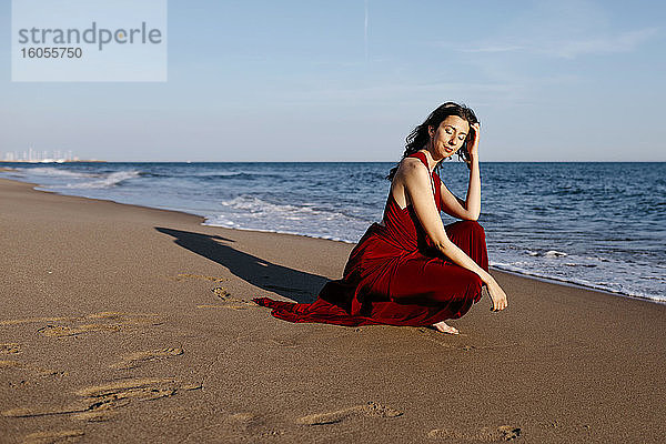 Zarte Frau in rotem Kleid sitzt am Strand und genießt die Sonne