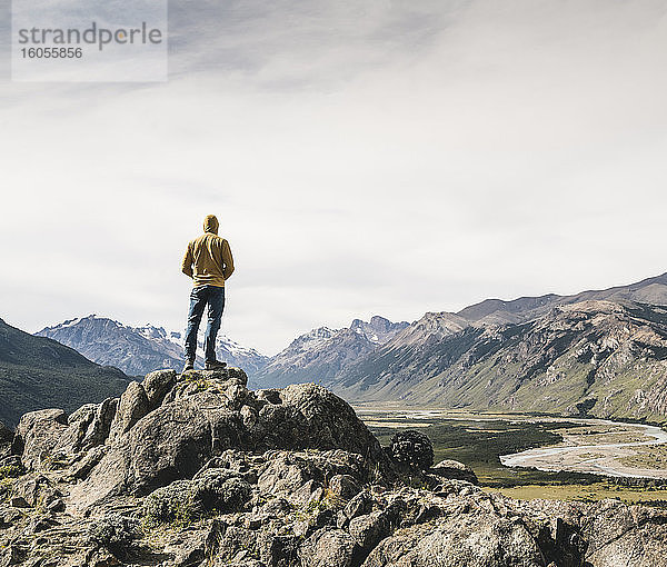 Älterer Mann mit Kapuze  der auf einem Felsen stehend die Berge gegen den Himmel betrachtet  Patagonien  Argentinien