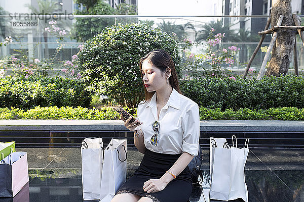 Junge Frau  die ein Smartphone benutzt  während sie mit Einkaufstüten auf dem Sitz in der Stadt sitzt