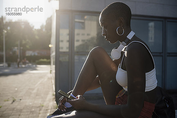 Junge Frau mit rasiertem Kopf  die ein Smartphone benutzt  während sie an einem sonnigen Tag in der Stadt sitzt