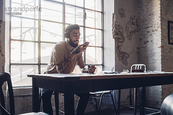 Kreativer junger Geschäftsmann mit Kopfhörern und Smartphone sitzt am Tisch im Loft-Büro