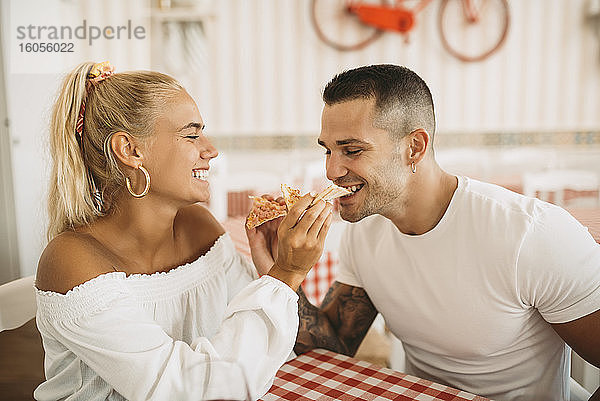 Fröhliche junge Frau füttert ihren Freund mit Pizza  während sie im Restaurant sitzt