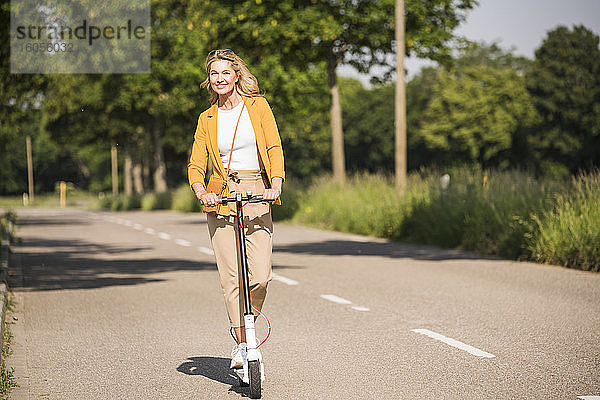Glückliche reife Frau fährt elektrischen Roller auf der Straße während des sonnigen Tages