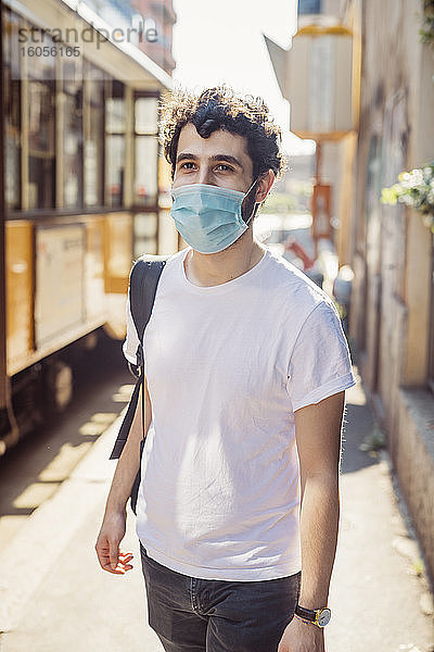 Junger Mann mit Maske  der an einem sonnigen Tag auf einer Straße in der Stadt spazieren geht