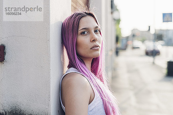 Porträt einer stilvollen jungen Frau mit rosa Haaren  die sich in der Stadt an eine Wand lehnt