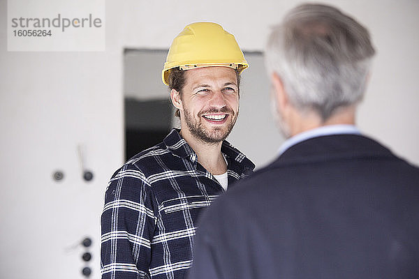 Lächelnder Bauarbeiter mit Schutzhelm im Gespräch mit einem Architekten beim Bau eines Hauses