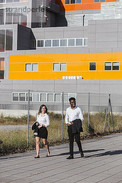 Lächelnde Kollegen  die an einem sonnigen Tag auf einem Fußweg vor einem modernen Gebäude spazieren gehen