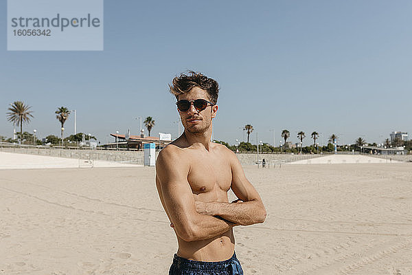 Hemdlose junge Frau mit Sonnenbrille und verschränkten Armen am Strand gegen den klaren Himmel stehend