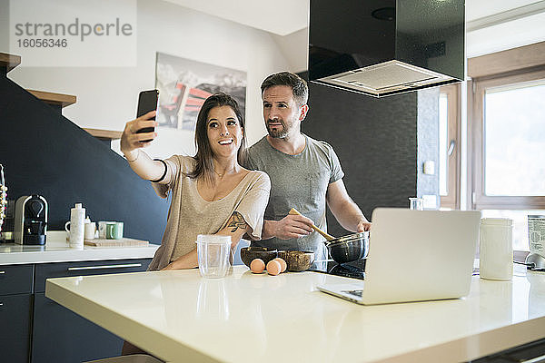 Lächelnde Frau nimmt Selfie mit Mann bereitet Essen auf Kücheninsel zu Hause
