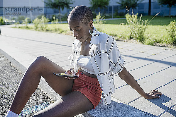 Junge Frau mit rasiertem Kopf  die ein Mobiltelefon benutzt  während sie auf einem Fußweg in der Stadt sitzt
