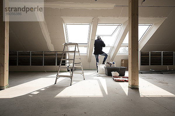 Männlicher Architekt  der über sein Smartphone spricht  während er am Fenster eines im Bau befindlichen Hauses steht