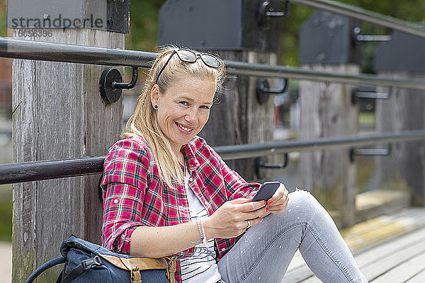 Lächelnde Frau im mittleren Erwachsenenalter  die ein Smartphone benutzt  während sie an der Reling auf dem Pier sitzt