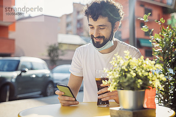 Lächelnder junger Mann mit Getränk und Smartphone auf dem Tisch  während er in einem Straßencafé sitzt