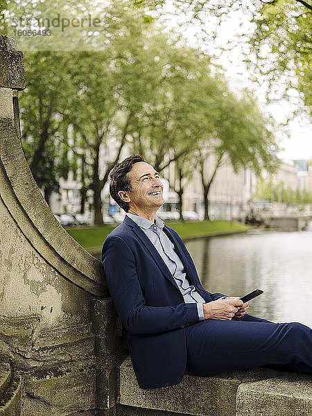 Glücklicher reifer Geschäftsmann mit Smartphone  der auf einer Brückenbrüstung sitzt