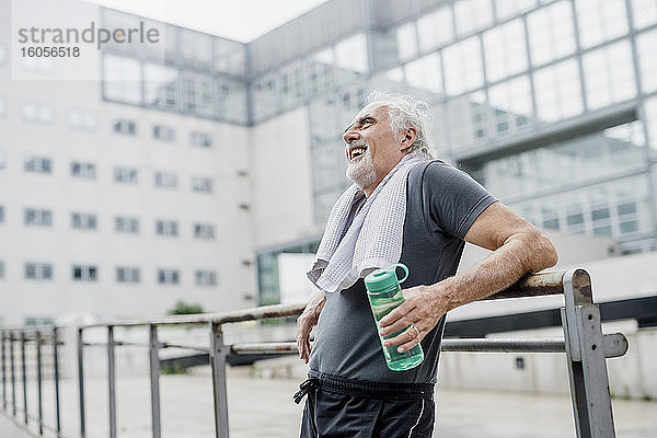 Fröhlicher älterer Mann  der eine Wasserflasche hält und wegschaut  während er am Geländer steht