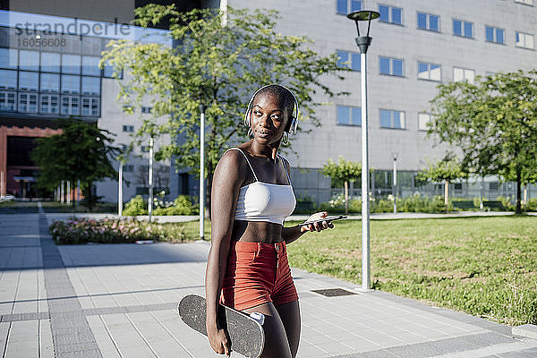 Junge Frau hält Handy und Skateboard und schaut weg  während sie auf einem Fußweg in der Stadt steht