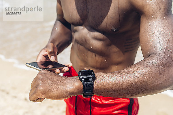 Sportlicher Mann mit Smartphone und Smartwatch am Strand