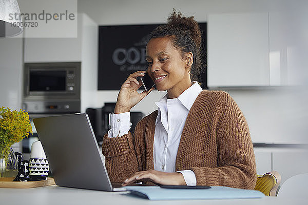 Lächelnde Geschäftsfrau am Telefon  die in der Küche sitzt und am Laptop arbeitet