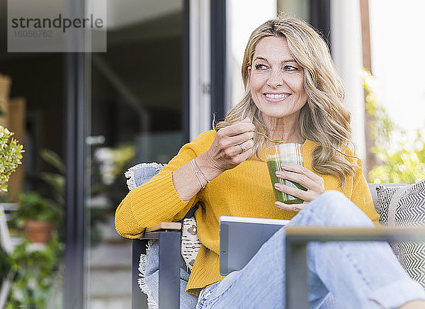 Porträt einer lächelnden reifen Frau  die mit einem digitalen Tablet auf einer Terrasse sitzt und einen grünen Smoothie genießt