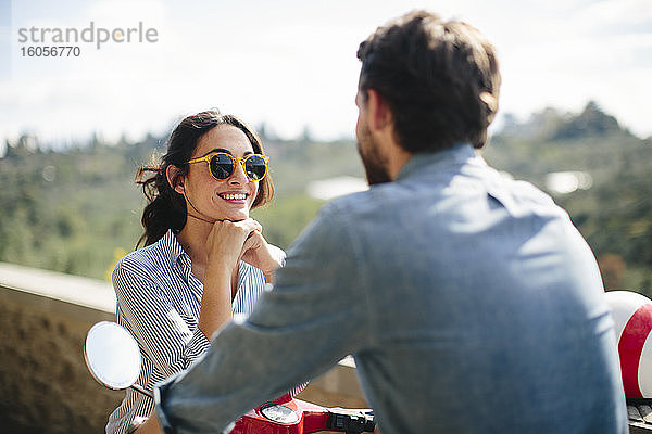 Lächelnde Frau mit Sonnenbrille im Gespräch mit ihrem Freund auf einer Vespa sitzend
