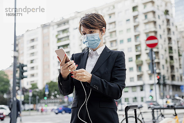 Geschäftsfrau mit Maske  die ein Smartphone und Kopfhörer hält  während sie in der Stadt steht