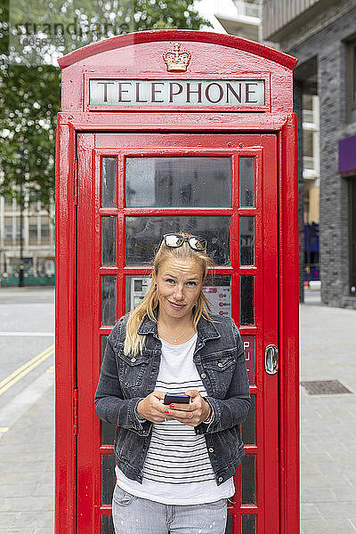 Mittlere erwachsene Frau  die ein Smartphone benutzt  während sie an einer Telefonzelle steht