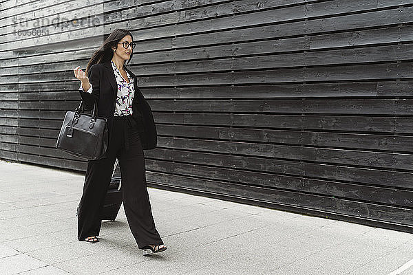 Selbstbewusste junge Geschäftsfrau mit Gepäck auf dem Fußweg an der schwarzen Wand
