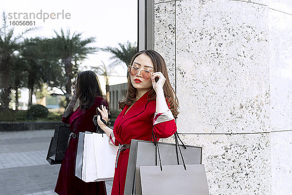Junge Frau mit Sonnenbrille trägt Einkaufstaschen  während sie an einem modernen Gebäude steht