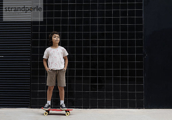 Junge mit Händen in den Taschen steht auf Skateboard gegen die Wand