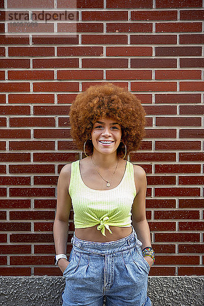 Lächelnde erwachsene Frau mit Afro-Haar  die gegen eine Mauer in der Stadt steht