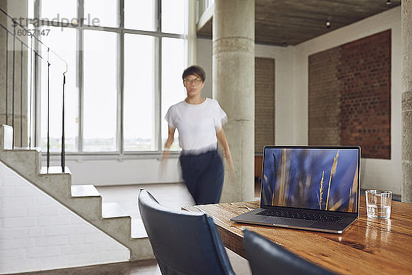 Laptop auf Holztisch in einer Loftwohnung mit Frau im Hintergrund
