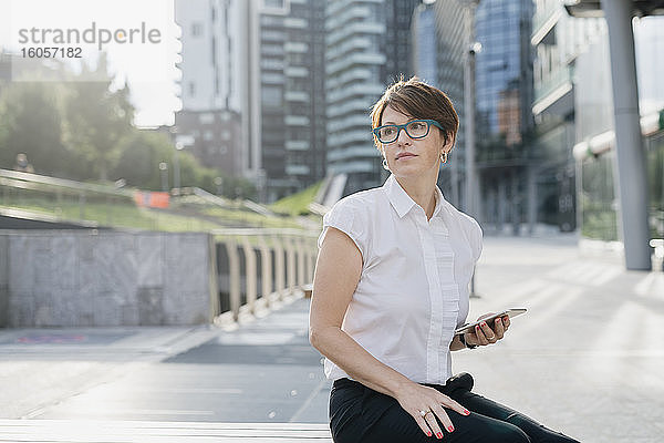 Nachdenkliche Geschäftsfrau hält ihr Smartphone in der Hand  während sie an einem Gebäude in der Stadt sitzt
