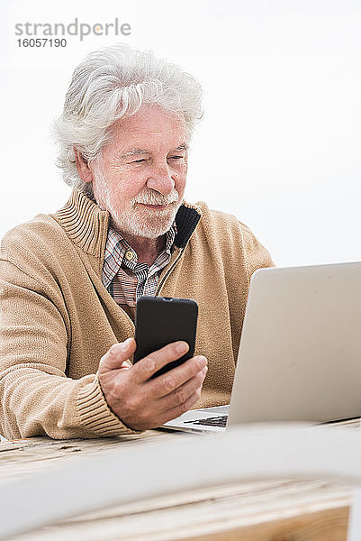 Älterer Mann hält ein Smartphone und benutzt einen Laptop am Tisch
