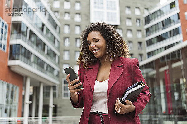 Lächelnde junge Geschäftsfrau  die ein Smartphone benutzt  während sie an einem Gebäude in der Stadt steht