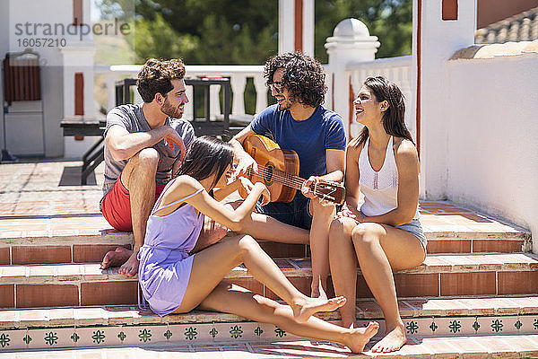Glückliche junge multiethnische Freunde  die sich mit einer Gitarre auf den Stufen einer Terrasse an einem sonnigen Tag vergnügen