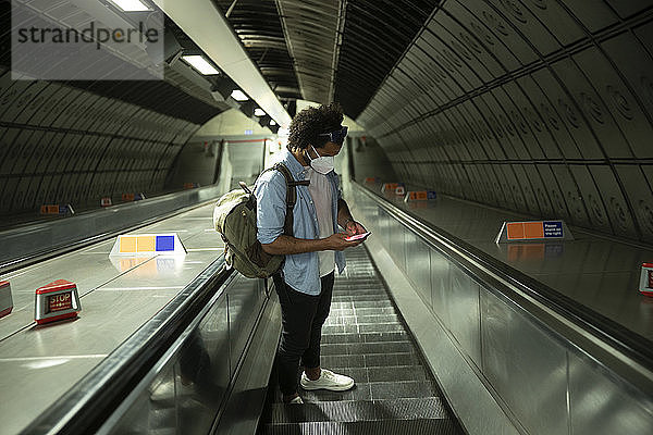 Mann mit Schutzmaske steht auf Rolltreppe und schaut auf sein Handy  London  UK
