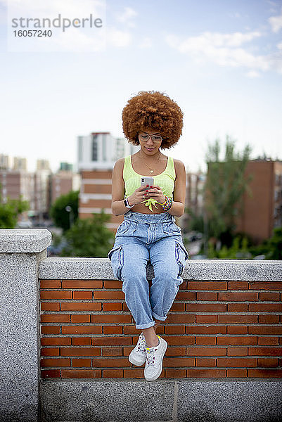 Frau mit Afrofrisur  die ihr Smartphone benutzt  während sie auf einer Stützmauer sitzt