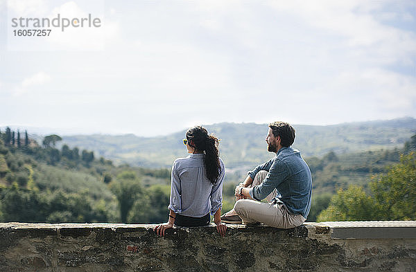 Paar betrachtet die Landschaft  während es auf einer Stützmauer gegen den Himmel sitzt