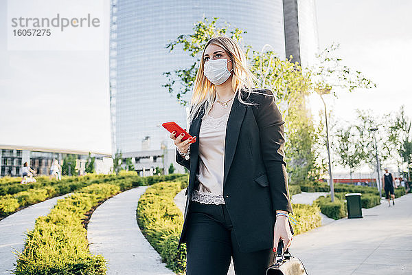 Geschäftsfrau mit Gesichtsmaske  die ein Smartphone hält  während sie auf einem Fußweg in der Stadt spazieren geht