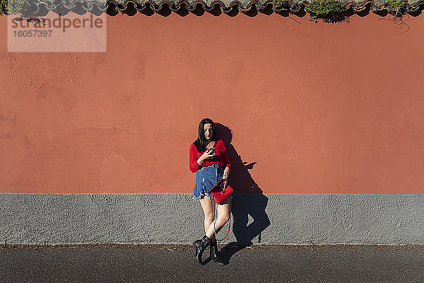 Junge Frau  die eine Handtasche hält und ein Smartphone benutzt  während sie sich an eine orangefarbene Wand in der Stadt lehnt