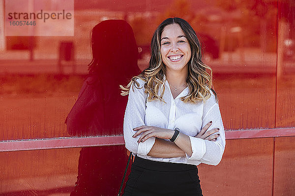 Lächelnde Geschäftsfrau mit verschränkten Armen vor einer roten Wand in der Stadt