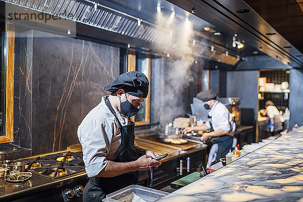 Koch mit Schutzmaske und Smartphone in der Restaurantküche