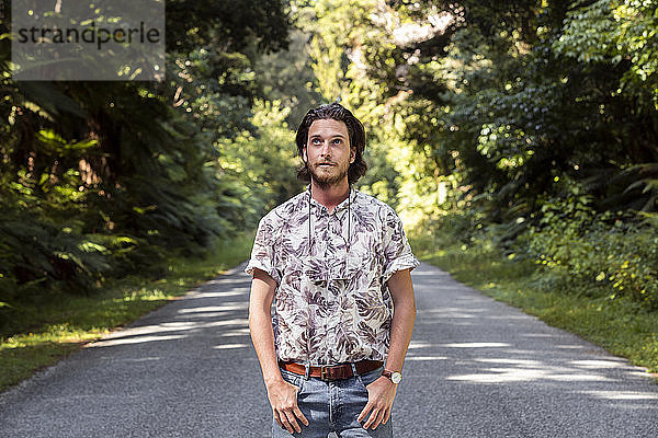 Nachdenklicher junger Mann steht auf der Straße inmitten von Bäumen im Wald